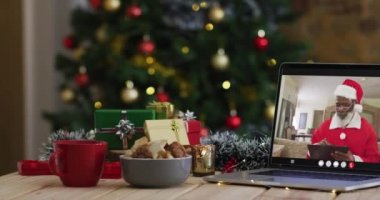 Kıdemli Afro-Amerikalı Noel Baba kostümlü adam dizüstü bilgisayarında Noel ağacıyla görüntülü arıyor. Noel, şenlik ve iletişim teknolojisi dijital kompozit video.