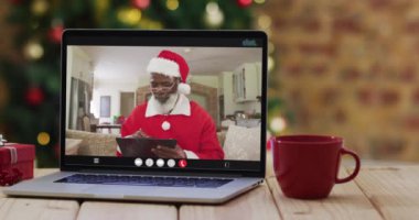 Kıdemli Afro-Amerikalı Noel Baba kostümlü adam dizüstü bilgisayarında Noel ağacıyla görüntülü arıyor. Noel, şenlik ve iletişim teknolojisi dijital kompozit video.