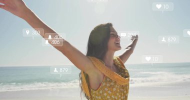 Sarı elbiseler giymiş, plajda kollarını kaldıran, yüzen sosyal medya simgeleriyle ayakta duran bir kadın. Eğlence, rahatlama, yaz, eğlence, canlılık, bağlantı, sükunet