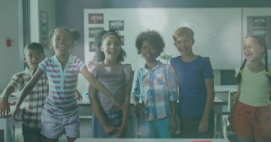 Sınıfta ayakta duran ve kollarını kaldıran gülümseyen sınıf arkadaşları, masa sandalyeleri beyaz tahta posterleriyle. Öğrenci, eğitim, öğrenim, takım çalışması, okul, keşif, gençlik