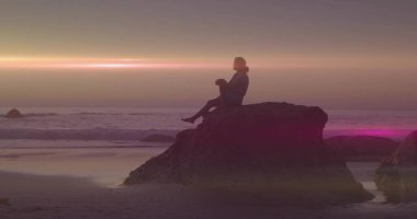 Uzun kollu bluzuyla oturan kadın ve şortuyla kumsaldaki alacakaranlıkta dalgaları seyrediyor. Yalnızlık, sükunet, sahil, gün batımı, yansıma, sükunet, doğa.