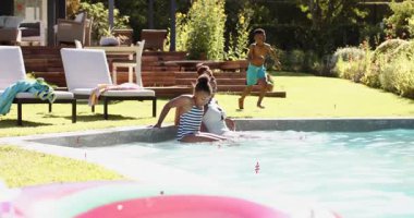 Anne ve kızı, oğullarının çimleri yarıştırıp babasının omuzlarına atladığı boş havuza su sıçratıyor. Aile, eğlence, boş zaman, açık hava, su, yaz zamanı, oyuncu.