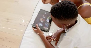 Çocuk yerde yatıp tablet aşırıyor düğümler açıyor ve kardeş gözlemi olarak teknoloji galerisinde geziniyor. Çocuklar, teknoloji, etkileşim, dijital, oyuncu, öğrenme, odaklanma