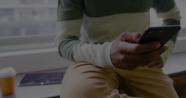 Looking at smartphone man in sweater on window ledge, with coffee cup and magazine, copy space. Urban, casual, modern, lifestyle, relaxation, interior, technology