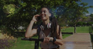 Dehşet verici bir kadın, e-scooter 'da dengede duruyor, parkta akıllı telefonla konuşuyor, kulaklık takıyor. Kentsel, yaşam tarzı, hareketli, boş zaman, açık hava, modern, enerjik