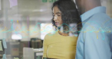 Ofiste bölümün yanında tablet tutan sarı bluzlu kadın, notlar, dalga formları. İşbirliği, yenilik, takım çalışması, dijital, iletişim, modernite, teknoloji