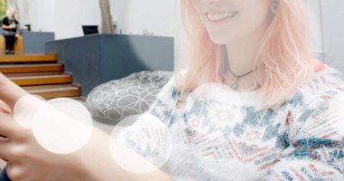 Akıllı telefonu tutan ve modern verandada puf üzerine oturmuş kolye takan gülümseyen kadın. Yaşam tarzı, gündelik, çağdaş, açık hava, sosyal, canlı, rahat