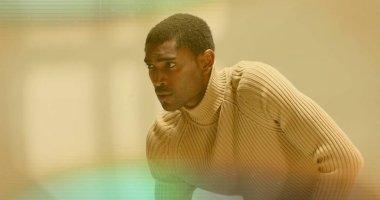 Minimalist stüdyo içinde bej çizgili kazağıyla sola bakan öne doğru eğilmiş bir adam. Portre, profesyonel, çağdaş, moda, zarafet, içe dönük, incelikli