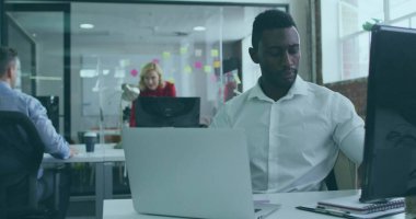 Afro-Amerikalı bir adam dizüstü bilgisayarla çalışıyor ve kahve fincanıyla açık planlı ofiste monitör ayarlıyor. İşbirliği, takım çalışması, modern, çalışma alanı, üretkenlik, iletişim, yenilik