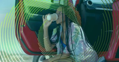 Kıyı şeridindeki açık kırmızı SUV 'nin yanında çiçekli örtüsü olan çömelmiş bir kadın kahve fincanından yudumluyor. Plaj giysisi, eğlence, seyahat, açık hava, yaşam tarzı, canlı, güneşli