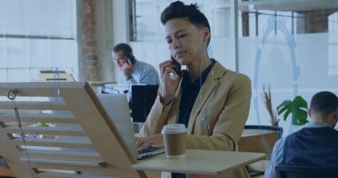 Akıllı telefonu tutan, spor ceketli Asyalı kadın ofisteki masada dizüstü bilgisayarda kahve fincanıyla yazı yazan. Eş zamanlı, profesyonel, modern, çalışma alanı, teknoloji, işbirliği, üretkenlik