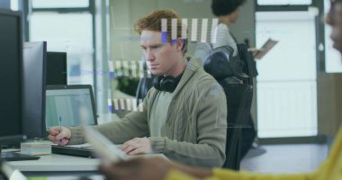 Ofis masasında çalışan kulaklıklı, dizüstü bilgisayarı, monitörü ve yapışkan notları olan bir adam. İşbirliği, üretkenlik, yenilik, modern, takım çalışması, teknoloji, çalışma alanı