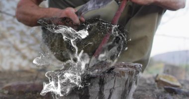 Kırsal kesimde, ahşap bir kütükte, ahşap keskinleştiren iş elbisesi içinde çömelmiş bir adam, ahşap parçalarıyla. Zanaatkârlık, zanaat, açık hava, engebeli, kırsal, faaliyet, kararlılık