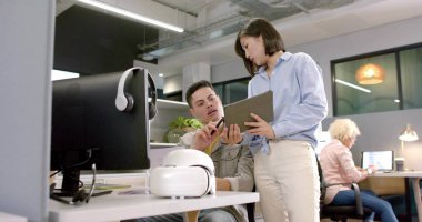 İş kıyafetleri içinde kadın iş arkadaşını gösteriyor. Erkek, açık ofiste tablet ve sanal gerçeklikle el kol hareketi yapıyor. İşbirliği, kulaklıklar, monitör, laptop, santral, bölünme, aydınlatma