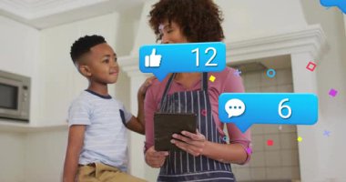 Anne tableti tıklatıyor, oğlana mutfakta rehberlik ediyor, yüzen sosyal medya rozetleri, nişanı güncelliyor. Aile, teknoloji, etkileşim, iletişim, yaşam tarzı, yerli, dijital