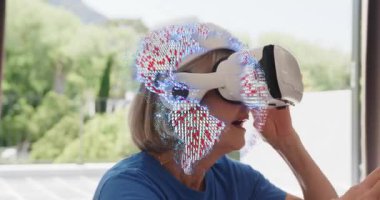 Masada dinlenen VR kulaklığa dikkat çeken yaşlı kadın kaldırma cihazı ve yüzen simgeleri keşfediyor. Son sınıf, teknoloji, yenilik, rahatlama, açık hava, modern, keşif