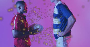 İki sporcu rugby topunu spor gösterisiyle yükselterek eğimli altın parçacıklar yayıyorlar. Sporcular, takım çalışması, enerji, dinamik, hareket, spor, yenilik