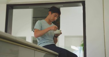 Koreli genç bir çocuk yemek yiyor mutfak tezgahının kenarındaki beyaz kaseden, saksı bitkisiyle yiyecek topluyor. Modern iç mekan, günlük kıyafetler, gençlik yaşam tarzı, modern ev, ev içi rahatlığı.