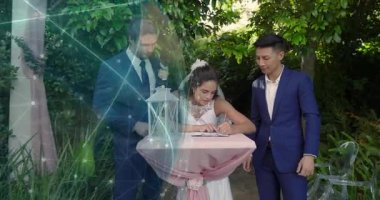 Gelin pembe düğün masasında kalem seçiyor, evlilik kaydını imzalıyor ve damat da destekliyor. Romantik, kutlama, zarafet, samimiyet, açık hava, neşeli, çiçekli