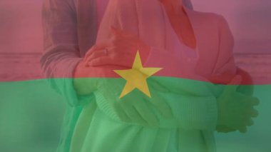 Burkina bayrağı solarken kıyı şeridini kucaklayan kıdemli çift, seyahat etmelerini engelliyor. Yaşlı, sarılma, plaj, deniz kenarı, ufuk, sakin, bulutlu
