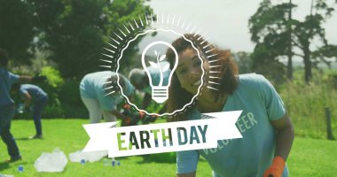 Turuncu eldivenli mavi tişörtlü kadın toplamak, parktaki çantalara şişeleri karıştırmak ve EARTHDAY örtüsü koymak. Gönüllü, temizlik, topluluk, çevre, güneş ışığı, takım çalışması, açık hava
