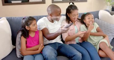 Uzaktan kumandaya basan adam, işaret eden kadın, ev teknolojisi için kanepede beliren Holo UI gibi gülen kızlar. Aile, televizyon, holografik, büyütme, etkileşim, rahat, oturma odası