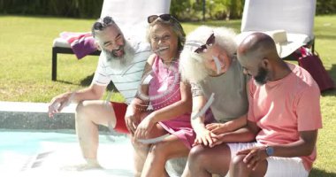 Dört arkadaş havuz kenarında oturuyor, kol saatini işaret ediyor, el hareketi teknoloji sarmalına neden oluyor. Havuz başı, arka bahçe, güneş ışığı, kahkaha, arkadaşlık, sohbet, gündelik