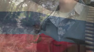 Asker, kamera kaldırırken göz kırpıyor. Kaskı, bandocuyu, askeri kimlik için üç renkli örtüyü gösteriyor. Asker, kamuflaj, yelek, orman, bayrak, açık hava, stok