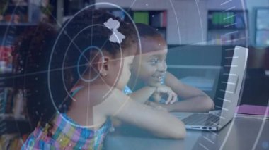 Kütüphanede dizüstü bilgisayara yaslanmış iki kız, ekranda HUD kaplama yapıyor, işaret ediyor, gülümsüyor ve öğreniyor. Eğitim, sınıf, öğrenciler, çocuklar, işbirliği, takım çalışması, teknoloji