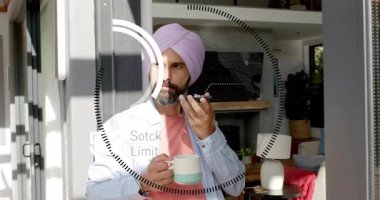 Adam yansıtılan HUD bisikletine cevap veriyor, telefonu kaldırıyor ve finans tablolarını izlerken konuşuyor. Uzaktan çalışma, kupa, cam kapı, güneş ışığı, ev, minimalist, odaklanma