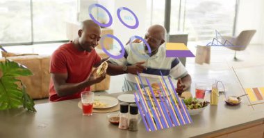 Yetişkin oğul ve büyük baba dirseklerini çarpıştırıyor, tabakları kaplayan animasyon yemek çizelgelerini paylaşırken yemeklerini paylaşıyorlar. Mutfak, açılış planı, modern, aydınlık, yemek, sohbet, kaynaşma