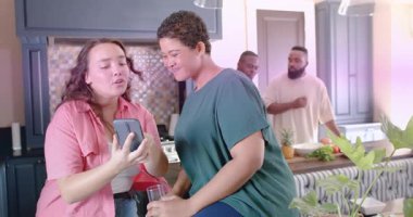 Telefon gösteren bir kadın, dinleme yapan bir arkadaş, sosyal bir mutfak, pembe çizgiler kesişen yüzler. Mobil, toplanma, arkadaşlar, canlı, samimi, mercek patlaması, üretim