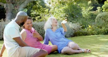 Tepedeki nesneye tepki veren iki kadın, kollarını kaldıran yaşlı bir kadın, sürüklenen teknoloji kutuları. Açık hava, park, çimen, üçlü, kahkaha, arkadaşlık, güneş ışığı