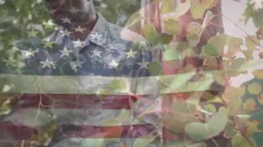 Ordu mensubu yaprakları itiyor, kamerayı kamufle ediyor, başlığı bayrak altında ayarlıyor. Kamuflaj, üniforma, açık hava, orman, yeşillik, gün ışığı, nokta