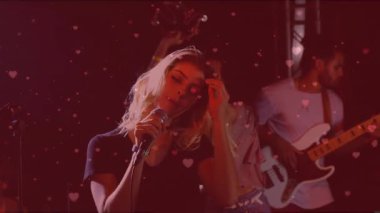 Baş şarkıcı mikrofona şarkı söylüyor ve saçlarını savuruyor. Müzik setinde havada süzülen kalpler başlatıyor. Sahne, grup, vokal, bas, davulcu, dansçı, kırmızı ışık