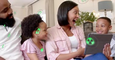 Anne tableti tıklatıyor, aileye koltukta rehberlik ediyor, yeşil simgeler sahne ve uygulama içeriğini örtüyor. Ebeveynler, çocuklar, oturma odası, aygıt, eşleştirme, etkileşim, parlaklık