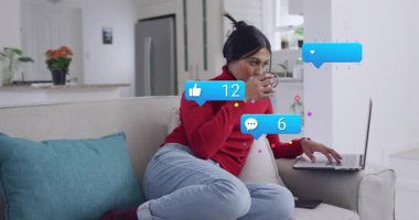 Kanepedeki kadın nişan kontrolü, bardak yudumlama, daktilo, dizüstü bilgisayarın etrafında yükselen sosyal tebligatlar. Koltuk, bilgisayar, içecek, ev, bitki, rahat, sakin