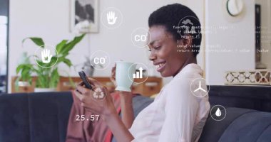 Kanepede kayan, bardağı yudumlayan, telefonu dinleyen ve CO2 ekosistem bilgilerini güncelleyen bir kadın. Akıllı telefon, veri, okumalar, santral, gün ışığı, ev, minimalizm