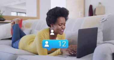 Kanepe dizüstü bilgisayarında yazan bir kadın, mavi rozetin yükselmesine neden oluyor, ikonalar sosyal ilişkileri artırıyor. Bildirimler, etkileşim, konfeti, oturma odası, güneş ışığı, rahatlama, odaklanma