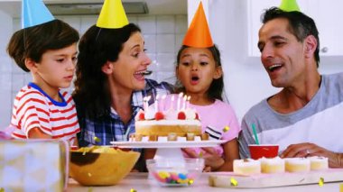 Masadaki aile, parti reklamı için kek mumları üfleyen tezahürat yapan kız, yüzlerin üzerinde konfeti animasyonu yapan kadın. Doğum günü, mutfak, kutlama, aileler, çocuklar, şapkalar, hediye