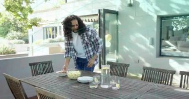 Ekoseli bir adam açık masada tabak düzenliyor, arkadaşları kapıya balonlarla girip yemek hazırlıyor. Alfresco, veranda, güneşli, toplanma, aile, servis, tabak