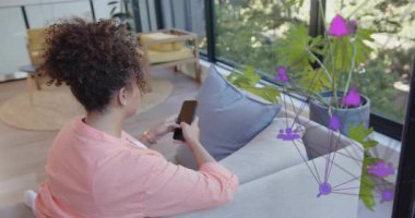 Oturan kadın telefonu dinliyor, cam boyunca mor ağ teknolojisine neden oluyor, telefonu kaldırıyor ve çağrıya katılıyor. Akıllı telefon, kadın, minimalist, ev, gün ışığı, bitkiler, yastıklar