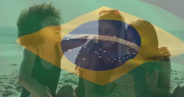 Brezilya bayrağının altında elinde telefonla gezen üç kadın gülüyor. Arkadaşlar, deniz kenarı, tatil, güneşli, mayo, güneş gözlüğü, akıllı telefon