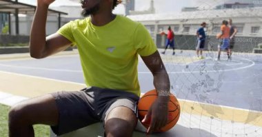 Yedekte oturan bir adam basketbol topunu tutuyor, teneffüste su içmek için içiyor, HUD mahkemeyi kaplıyor. Açık hava, şehir, atlet, hidrasyon, top, takım arkadaşları, güneşli