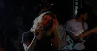 Koro solisti saç kırbaçlıyor ve sahnede mikrofon şakıyarak şarkı söylüyor. Animasyon ışıkları vurgulanıyor. Performans, konser, orkestra, vokal, gitarist, basçı, davulcu