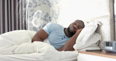 Yatakta uyanan bir adam, beyaz bir girdap beliriyor, bilek kontrolüne, oturmaya, sağlık için esnemeye neden oluyor. Gün ışığı, sabah, esneme, dinginlik, sağlık, yatak odası, yatak başlığı