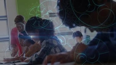 Öğretmen öğrencilerin eğilip çalışmalarına destek olurken dişli halkaları eğitimi süpürüyor. Sınıflar, öğrenciler, öğrenciler, masalar, gün ışığı, pencere, işbirliği