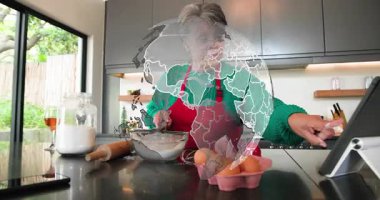 Kıdemli kadın tablete danışmanlık yapıyor ve mutfakta un eleme yapıyor. Tarifi çırpmaya rehberlik eden animasyon adımlar atıyor. Ada, yemek, malzemeler, pişirme, arkadaşlık, güneş ışığı, önlük.