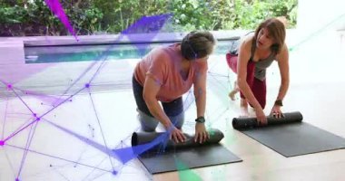 İki kadın verandada kafa sallıyor ve hasırları açıyor. AR ise renklendirici spor paspasları örtüyor. Spor, açık hava, esneme, arkadaşlar, bahçe, havuz, gün ışığı.