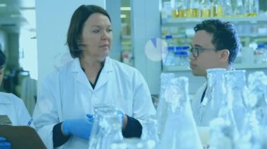 Laboratuvarda konuşan ve el kol hareketi yapan kıdemli bilim adamı, şişelerin üzerinde mor çizelgeler başlatıyor, rehberlik ekibi. Araştırmacı, takım çalışması, laboratuvar, bilim, cam eşyalar, deney tüpleri, kaplama